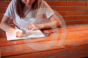 The girl wearing a white shirt sitting on a wooden table and writing a book. There is sunshine in the hands of a young woman.