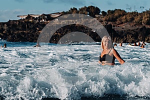 A girl with wet hair and a black swimsuit stands in the white foam of sea water on the island of Tenerife, around a wave with