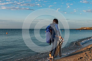 A girl in a wetsuit with flippers ih her hands and dry bag walking along the beach