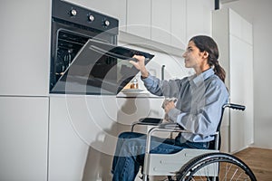 A girl on a wheelchair opening oven in the kitchen while cooking soemthing