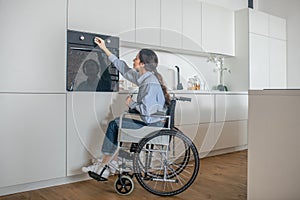 A girl on a wheelchair opening oven in the kitchen while cooking soemthing