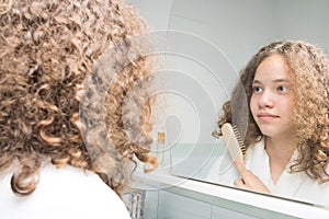 Girl in a white bathrobe in the bathroom standing in front of the mirror combing her curly hair