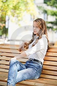 A girl in a white blouse and blue jeans sits on a wooden bench in autumn Park square