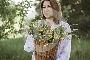 A girl in a white blouse holds a wicker basket with a bouquet of wild flowers. Summer walk in the field