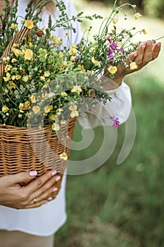 A girl in a white blouse holds a wicker basket with a bouquet of wild flowers. Summer walk in the field. midsection