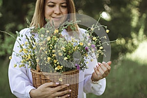 A girl in a white blouse holds a wicker basket with a bouquet of wild flowers. Summer walk in the field. midsection