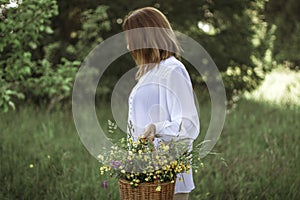 A girl in a white blouse holds a wicker basket with a bouquet of wild flowers. Summer walk in the field. midsection