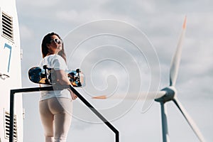 A girl in white clothes and glasses with a skate in her hands is photographed near large wind turbines in a field with trees.