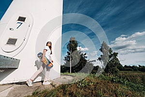 A girl in white clothes and glasses with a skate in her hands is photographed near large wind turbines in a field with trees.