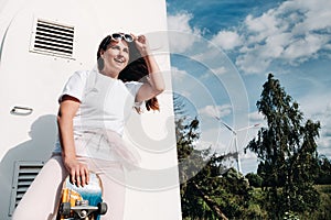 A girl in white clothes and glasses with a skate in her hands is photographed near large wind turbines in a field with trees.