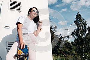 A girl in white clothes and glasses with a skate in her hands is photographed near large wind turbines in a field with trees.