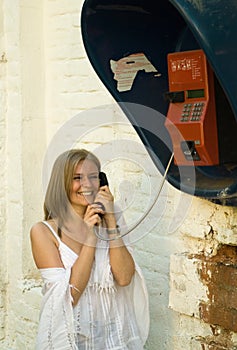 A girl in white clothes talks cheerfully on a street payphone.