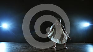 A girl in a white dress dances contemporary making rotations on the stage with smoke in spotlights.