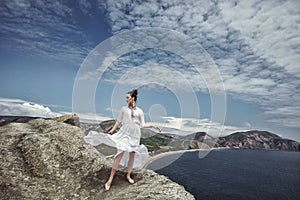 A girl in a white dress and decorations stands on a cliff against the background of the sea and mountains, her dress is spread fro