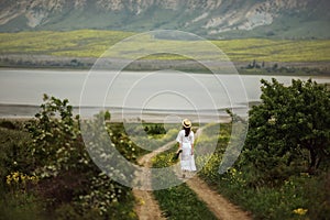 A girl in a white dress, in an elegant hat with a bouquet of yellow field flowers walks down a rural road in the middle of green m