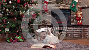 A girl in a white dress by the fireplace under the Christmas tree is lying on her stomach and reading a book. Festive