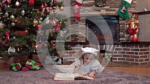 A girl in a white dress by the fireplace under the Christmas tree is lying on her stomach and reading a book. Festive