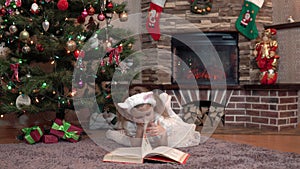 A girl in a white dress by the fireplace under the Christmas tree is lying on her stomach and reading a book. Festive atmosphere