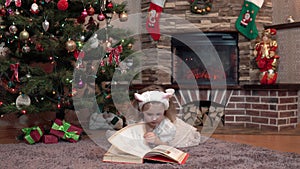 A girl in a white dress by the fireplace under the Christmas tree is lying on her stomach and reading a book. Festive atmosphere