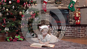 A girl in a white dress by the fireplace under the Christmas tree is lying on her stomach and reading a book. Festive atmosphere