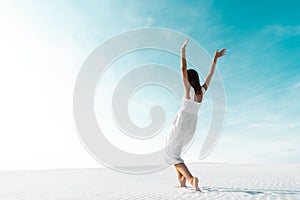 Girl in white dress with hands in air on sandy beach with blue sky
