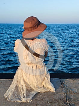 girl in a white dress and hat sits on the pier against the backdrop of the sea horizon, rear view