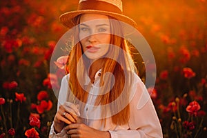 a girl in a white dress and hat stands in a field with poppies at sunset and holds a poppy flower in her hand