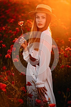 a girl in a white dress and hat stands in a field with poppies at sunset and holds a poppy flower in her hand