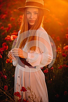 a girl in a white dress and hat stands in a field with poppies at sunset and holds a poppy flower in her hand