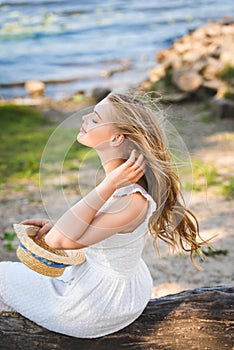 Girl in white dress holding straw hat while sitting wooden trunk with closed eyes
