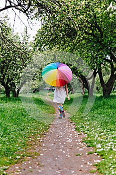 Girl in a white dress jumping in the blooming garden with colorful rainbow-umbrella. Spring, outdoors.