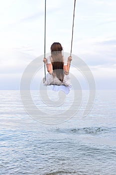 Girl in white dress is riding swing above water. Vertical frame. Back view. Background of blue sky horizon and sea