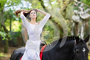 A girl in a white dress sits on a horse and straightens her hair. Outdoors.