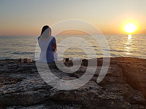 A girl in a white dress sitting on the beach on a stone and watching the sunset. The yellow-orange sun hangs on the horizon