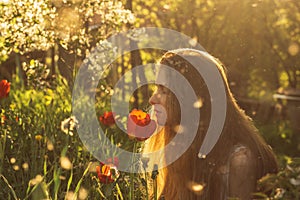 Girl in white dress smelling tulip in sunset among fluff, dandelions and cherry flowers