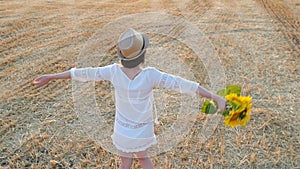 A girl in a white dress and a straw hat is holding a bouquet of flowers from a sunflower and is spinning. Young woman in