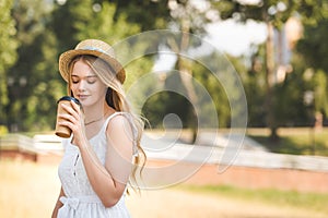 Girl in white dress and straw hat holding paper cup and drinking coffee with closed eyes