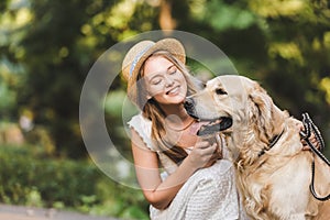 Girl in white dress and straw sitting near golden retriever and smiling while looking at dog