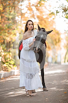 A girl in a white dress is walking with a horse in an autumn park.