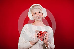 Girl in white fur headphones in a warm sweater on a red background. A blonde woman holding a heart in her hands. Photo in the