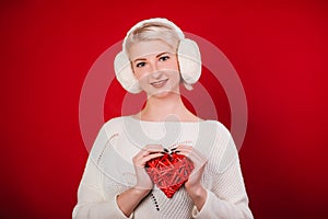 Girl in white fur headphones in a warm sweater on a red background. A blonde woman holding a heart in her hands. Photo in the