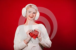 Girl in white fur headphones in a warm sweater on a red background. A blonde woman holding a heart in her hands. Photo in the