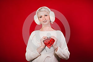 Girl in white fur headphones in a warm sweater on a red background. A blonde woman holding a heart in her hands. Photo in the