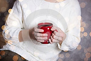 A girl in a white knitted sweater holds a red mug with tea in her hands. Copy space