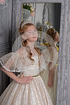 A girl in a white lace dress with a diadem in her hair, a young aristocrat, a ball gown, trying on a dress in front of a mirror