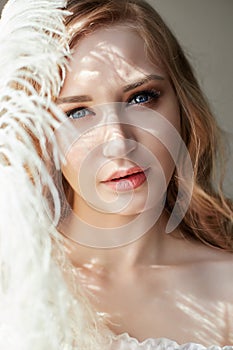 Girl in white light dress and curly hair with big feather near her face, portrait woman with feather, purity and innocence. Curly