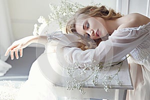 Girl white light dress and curly hair, portrait of woman with flowers at home near the window, purity and innocence. Curly blonde