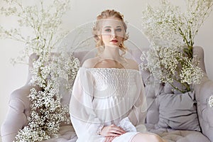 Girl white light dress and curly hair, portrait of woman with flowers at home near the window, purity and innocence. Curly blonde