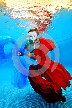 The girl in the white mask swims underwater with red and blue fabrics on the background of the sunset and looking at the camera.