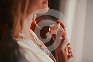 A girl in a white satin dressing gown with a neckline and a manicure french holds a bottle with perfume and splashes on her wrist